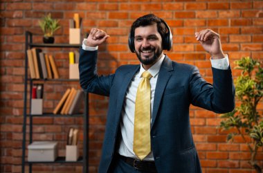 Kulaklık takıp neşeyle dans eden Hintli işadamı, başarıyı kutluyor ve modern ofiste neşeli müzik molası veriyor. Resmi kıyafetlerle rahat ofiste gülümsüyor ve ritimle hareket ediyor.