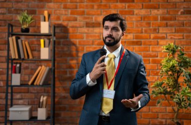 Hintli işadamı mikrofona konuşurken samimi modern ofiste resmi bir konuşma yapıyor. Asya CEO 'su ya da takım elbiseli ve sakallı girişimci kırmızı tuğla duvara yaslanmış duruyor.