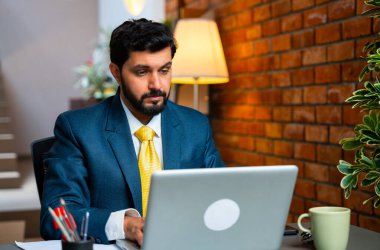 Laptop üzerinde çalışan ve derin düşünen Hintli işadamı, tatilde ofis penceresinden dışarı bakarken terasotta tuğlalı duvarlı, samimi kurumsal çalışma alanında resmi kıyafetler giyerken geleceği hayal ediyor.