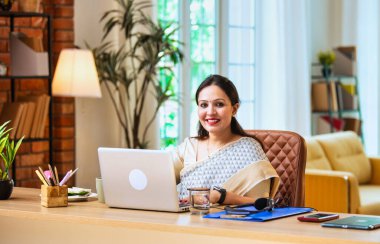 Kameraya bakıyor ve güvenle gülümsüyor. Etnik sari giymiş Hintli genç iş kadını ya da kadın kurumsal kadın portresi rahat ofisinde dizüstü bilgisayarla oturuyor.