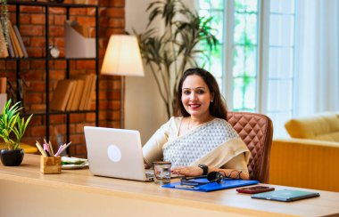 Kameraya bakıyor ve güvenle gülümsüyor. Etnik sari giymiş Hintli genç iş kadını ya da kadın kurumsal kadın portresi rahat ofisinde dizüstü bilgisayarla oturuyor.