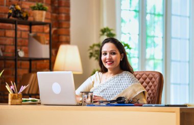 Kameraya bakıyor ve güvenle gülümsüyor. Etnik sari giymiş Hintli genç iş kadını ya da kadın kurumsal kadın portresi rahat ofisinde dizüstü bilgisayarla oturuyor.