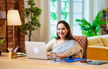 Kameraya bakıyor ve güvenle gülümsüyor. Etnik sari giymiş Hintli genç iş kadını ya da kadın kurumsal kadın portresi rahat ofisinde dizüstü bilgisayarla oturuyor.