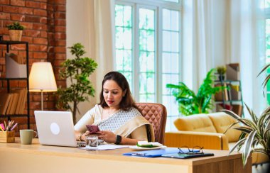 Ofis masasında çalışırken akıllı telefon kullanan Hintli kadın girişimci, iş saatlerinde cep telefonunda e-posta ya da fotoğraf kontrolü yapıyor, modern şirket ortamında dizüstü bilgisayarla birden fazla iş yapıyor.
