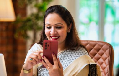 Ofis masasında çalışırken akıllı telefon kullanan Hintli kadın girişimci, iş saatlerinde cep telefonunda e-posta ya da fotoğraf kontrolü yapıyor, modern şirket ortamında dizüstü bilgisayarla birden fazla iş yapıyor.