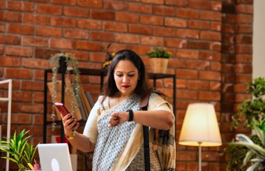 Akıllı telefondan taksi tutarken bilek saatini kontrol ediyorum. Endişeli Hintli genç iş kadını ya da etnik sari giymiş kurumsal bir bayan toplantı için gitmeden önce rahat modern ofis yerinde duruyor.