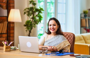 Kameraya bakıyor ve güvenle gülümsüyor. Etnik sari giymiş Hintli genç iş kadını ya da kadın kurumsal kadın portresi rahat ofisinde dizüstü bilgisayarla oturuyor.