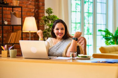 Dizüstü bilgisayarla çalıştıktan sonra zaferi kutlayan, etnik sarili Hintli genç iş kadını, rahat ofis masasında otururken zafer yumruğunu neşeyle gösterdi, kadın girişimci kariyer kilometre taşına ulaştı