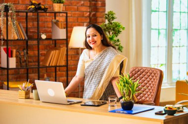 Etnik saride çalışan Hintli genç işkadını ya da kurumsal kadın işveren, ofis masasında dizüstü bilgisayarın yanında rahat çalışma alanındaki kameraya bakarak gülümsüyor.