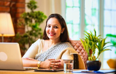 Kameraya bakıyor ve güvenle gülümsüyor. Etnik sari giymiş Hintli genç iş kadını ya da kadın kurumsal kadın portresi rahat ofisinde dizüstü bilgisayarla oturuyor.