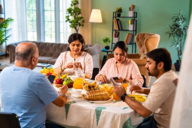 Birlikte kahvaltı yapıyorlar, Hintli üst düzey ebeveynler ve genç yetişkin kız ve erkek çocuklar rahat bir evde, modern yemek masasında sessizce oturuyorlar, akıllı telefonlar kullanıyorlar, sabah yemeği sırasında dijital olarak dikkatleri dağılıyorlar.