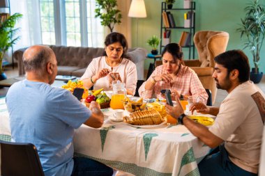 Birlikte kahvaltı yapıyorlar, Hintli üst düzey ebeveynler ve genç yetişkin kız ve erkek çocuklar rahat bir evde, modern yemek masasında sessizce oturuyorlar, akıllı telefonlar kullanıyorlar, sabah yemeği sırasında dijital olarak dikkatleri dağılıyorlar.