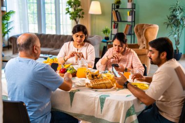 Birlikte kahvaltı yapıyorlar, Hintli üst düzey ebeveynler ve genç yetişkin kız ve erkek çocuklar rahat bir evde, modern yemek masasında sessizce oturuyorlar, akıllı telefonlar kullanıyorlar, sabah yemeği sırasında dijital olarak dikkatleri dağılıyorlar.