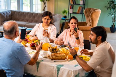 Birlikte kahvaltı yapıyorlar, Hintli üst düzey ebeveynler ve genç yetişkin kız ve erkek çocuklar rahat bir evde, modern yemek masasında sessizce oturuyorlar, akıllı telefonlar kullanıyorlar, sabah yemeği sırasında dijital olarak dikkatleri dağılıyorlar.