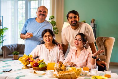 Gülümseyerek kameraya bakıyorum, neşeli Hintli aile, kıdemli ebeveynler ve genç yetişkin kız ve erkek çocuklar birlikte kahvaltı masasında oturuyorlar mutlu bir sabah anlarında.
