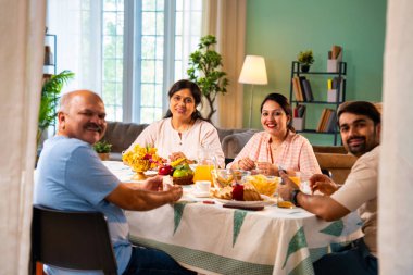 Gülümseyerek kameraya bakıyorum, neşeli Hintli aile, kıdemli ebeveynler ve genç yetişkin kız ve erkek çocuklar birlikte kahvaltı masasında oturuyorlar mutlu bir sabah anlarında.