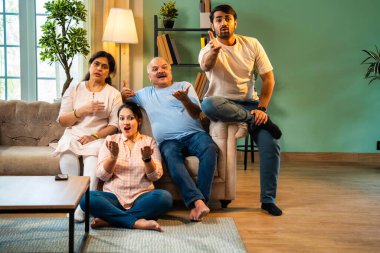 Kameraya şaşırmış, mutlu Hintli aile, yaşlı aileler ve genç yetişkin kız ve erkek çocuk modern oturma odasındaki koltukta oturmuş evdeki şaşırtıcı derecede komik bir şeye tepki gösteriyorlar.