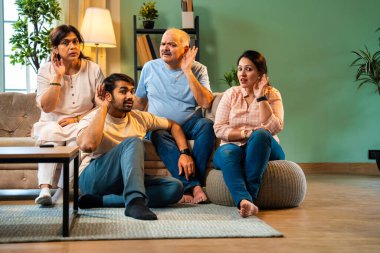 Kameraya bakarken kulakları, genç yetişkin bir oğlu ve kızı olan Hintli bir aile rahat modern oturma odasında oturup dedikodu ya da gizli şeyler dinliyorlar.