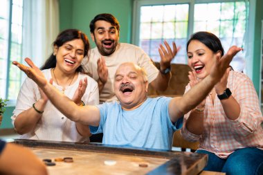 Birlikte carom ya da carrom masa üstü oyunu oynayarak, dört kişilik Hintli aile, yaşlı ebeveynler ve genç yetişkin bir çocuk ve kız evdeki boş zamanlarında rahat oturma odasında eğleniyorlar.