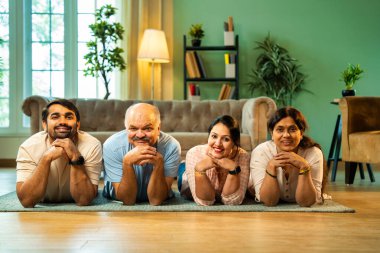 Ellerinin üzerinde yerde uzanmış, kameraya gülümsüyor, yetişkin bir oğlu ve kızı olan Hintli ebeveynler sıcak ev içlerinde birlikte poz veriyor, samimiyet, yakınlık ve birliktelik gösteriyorlar.