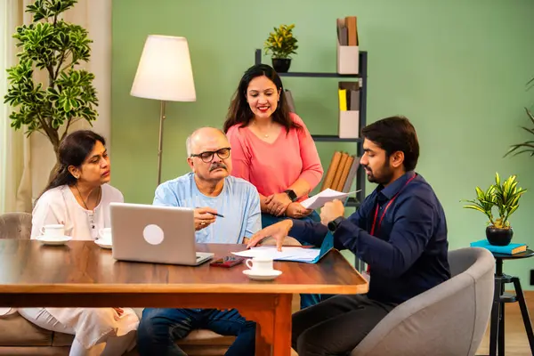 Hintli kıdemli bir adam, genç yönetici olarak belgeyi imzalarken ona yol gösteriyor. Rahat modern bir evde eşi ve kızının yanında oturuyor. Vasiyeti, yasal belgeleri, finans veya sigorta danışmanlığını sembolize ediyor.