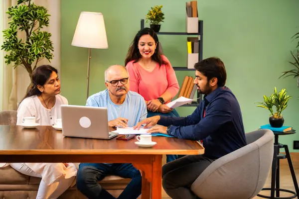 Hintli kıdemli bir adam, genç yönetici olarak belgeyi imzalarken ona yol gösteriyor. Rahat modern bir evde eşi ve kızının yanında oturuyor. Vasiyeti, yasal belgeleri, finans veya sigorta danışmanlığını sembolize ediyor.