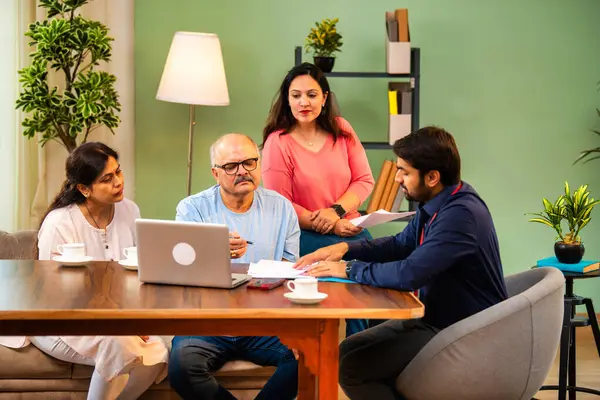 Hintli kıdemli bir adam, genç yönetici olarak belgeyi imzalarken ona yol gösteriyor. Rahat modern bir evde eşi ve kızının yanında oturuyor. Vasiyeti, yasal belgeleri, finans veya sigorta danışmanlığını sembolize ediyor.