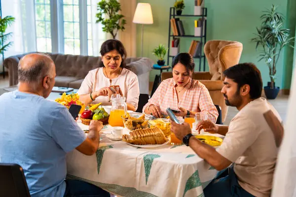 Birlikte kahvaltı yapıyorlar, Hintli üst düzey ebeveynler ve genç yetişkin kız ve erkek çocuklar rahat bir evde, modern yemek masasında sessizce oturuyorlar, akıllı telefonlar kullanıyorlar, sabah yemeği sırasında dijital olarak dikkatleri dağılıyorlar.