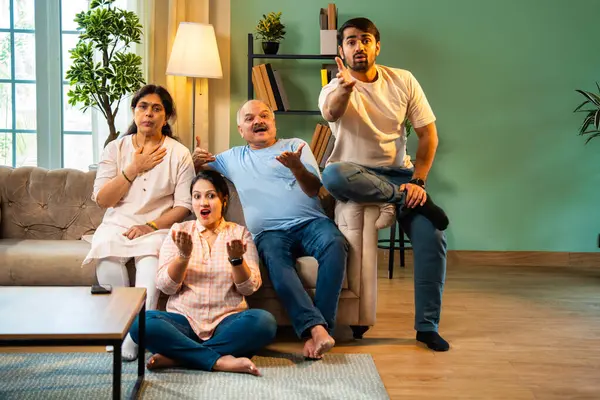 Kameraya şaşırmış, mutlu Hintli aile, yaşlı aileler ve genç yetişkin kız ve erkek çocuk modern oturma odasındaki koltukta oturmuş evdeki şaşırtıcı derecede komik bir şeye tepki gösteriyorlar.