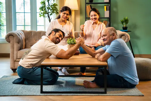 Hindistan 'ın kıdemli babasıyla genç yetişkin oğlu arasında bilek güreşi eğlencesi. Kıdemli eş ve genç kız gülüyor ve tezahürat yapıyorlar. Evde arkadaşça eğleniyorlar.