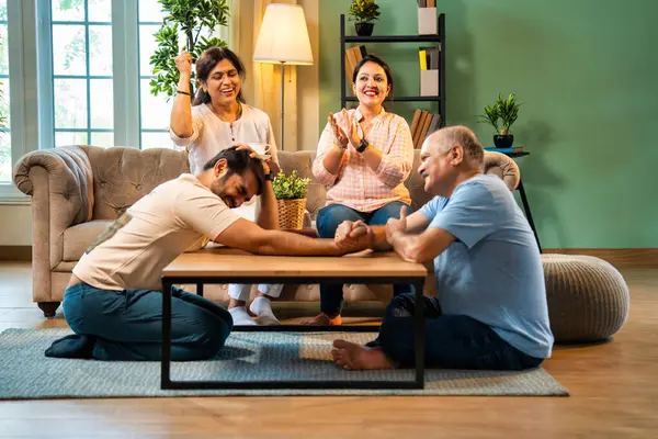 Hindistan 'ın kıdemli babasıyla genç yetişkin oğlu arasında bilek güreşi eğlencesi. Kıdemli eş ve genç kız gülüyor ve tezahürat yapıyorlar. Evde arkadaşça eğleniyorlar.