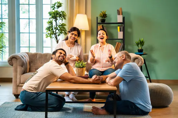 Hindistan 'ın kıdemli babasıyla genç yetişkin oğlu arasında bilek güreşi eğlencesi. Kıdemli eş ve genç kız gülüyor ve tezahürat yapıyorlar. Evde arkadaşça eğleniyorlar.