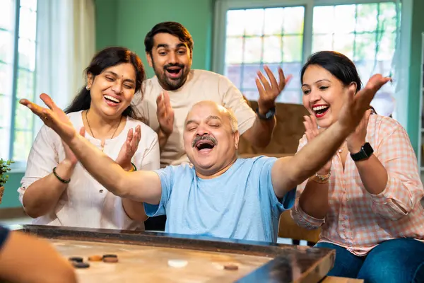 Birlikte carom ya da carrom masa üstü oyunu oynayarak, dört kişilik Hintli aile, yaşlı ebeveynler ve genç yetişkin bir çocuk ve kız evdeki boş zamanlarında rahat oturma odasında eğleniyorlar.