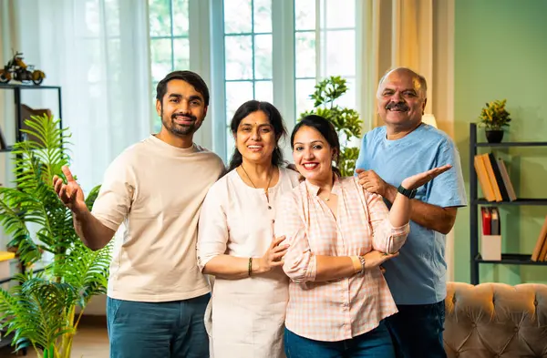 Kameraya bakıp gülümseyen, dört çocuklu Hintli aile, kıdemli ebeveynler ve genç yetişkin kız ve erkek çocuklar modern oturma odasında birlikte poz veriyorlar. Mutlu ve birlik olmuş görünüyorlar.