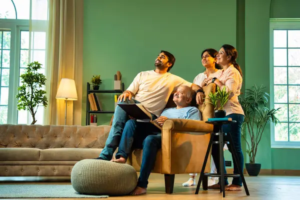 Yan yana baktığımızda, dört çocuklu Hintli aile, büyük ebeveynler ve küçük yetişkin çocuklar kanat sandalyesinde babalarının etrafında oturuyorlar, evde mutlu ifadelerle eski fotoğraf albümlerinin keyfini çıkarıyorlar.