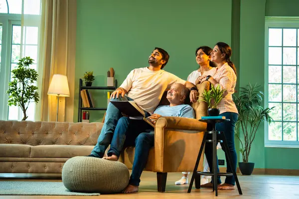 Yan yana baktığımızda, dört çocuklu Hintli aile, büyük ebeveynler ve küçük yetişkin çocuklar kanat sandalyesinde babalarının etrafında oturuyorlar, evde mutlu ifadelerle eski fotoğraf albümlerinin keyfini çıkarıyorlar.