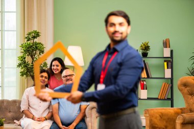 Evin önünü tutan Hintli genç bankacı ya da finans yöneticisi, arka plandaki koltukta oturan mutlu aile ev kredisi, finans ya da emlak kavramını temsil ederken gülümsüyor.