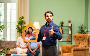 Evin önünü tutan Hintli genç bankacı ya da finans yöneticisi, arka plandaki koltukta oturan mutlu aile ev kredisi, finans ya da emlak kavramını temsil ederken gülümsüyor.