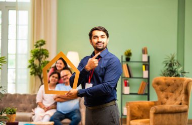 Evin önünü tutan Hintli genç bankacı ya da finans yöneticisi, arka plandaki koltukta oturan mutlu aile ev kredisi, finans ya da emlak kavramını temsil ederken gülümsüyor.