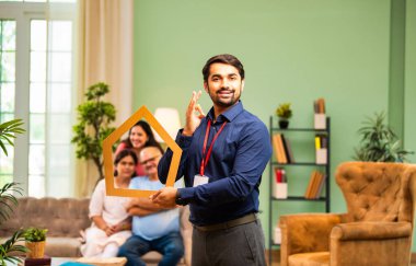 Evin önünü tutan Hintli genç bankacı ya da finans yöneticisi, arka plandaki koltukta oturan mutlu aile ev kredisi, finans ya da emlak kavramını temsil ederken gülümsüyor.