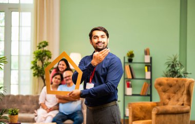Evin önünü tutan Hintli genç bankacı ya da finans yöneticisi, arka plandaki koltukta oturan mutlu aile ev kredisi, finans ya da emlak kavramını temsil ederken gülümsüyor.