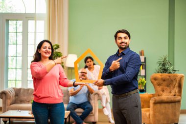 Koltuğun arkasında yaşlı ebeveynlerin oturduğu ev maketi, gülümseyen Hintli çift emlak hayalleri, mülk sahipliğini, ev kredisi finansmanını, aile yatırım planlamasını sembolize ediyor.