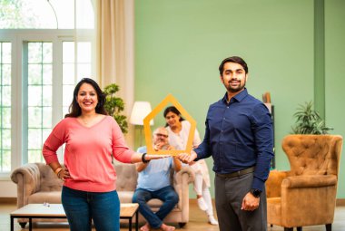 Koltuğun arkasında yaşlı ebeveynlerin oturduğu ev maketi, gülümseyen Hintli çift emlak hayalleri, mülk sahipliğini, ev kredisi finansmanını, aile yatırım planlamasını sembolize ediyor.