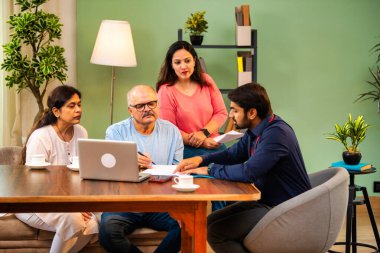 Hintli kıdemli bir adam, genç yönetici olarak belgeyi imzalarken ona yol gösteriyor. Rahat modern bir evde eşi ve kızının yanında oturuyor. Vasiyeti, yasal belgeleri, finans veya sigorta danışmanlığını sembolize ediyor.