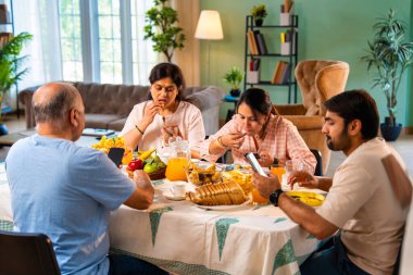 Birlikte kahvaltı yapıyorlar, Hintli üst düzey ebeveynler ve genç yetişkin kız ve erkek çocuklar rahat bir evde, modern yemek masasında sessizce oturuyorlar, akıllı telefonlar kullanıyorlar, sabah yemeği sırasında dijital olarak dikkatleri dağılıyorlar.