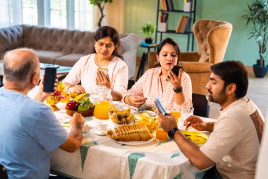 Birlikte kahvaltı yapıyorlar, Hintli üst düzey ebeveynler ve genç yetişkin kız ve erkek çocuklar rahat bir evde, modern yemek masasında sessizce oturuyorlar, akıllı telefonlar kullanıyorlar, sabah yemeği sırasında dijital olarak dikkatleri dağılıyorlar.