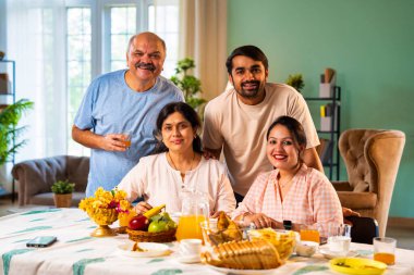 Gülümseyerek kameraya bakıyorum, neşeli Hintli aile, kıdemli ebeveynler ve genç yetişkin kız ve erkek çocuklar birlikte kahvaltı masasında oturuyorlar mutlu bir sabah anlarında.