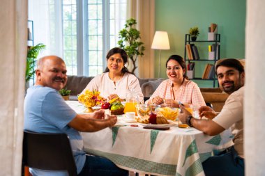 Gülümseyerek kameraya bakıyorum, neşeli Hintli aile, kıdemli ebeveynler ve genç yetişkin kız ve erkek çocuklar birlikte kahvaltı masasında oturuyorlar mutlu bir sabah anlarında.