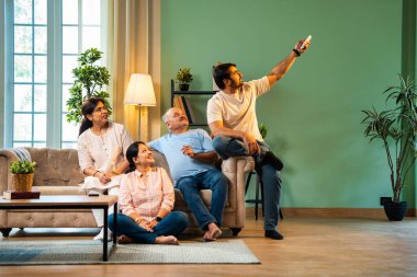 Sıcak ve terli hisseden Hintli aile, yaşlı ebeveynler ve genç yetişkin kız ve erkek çocuk modern oturma odasındaki koltukta oturuyorlar, kız klimayı uzaktan kumandayla açıyor, hepsi rahatlamış hissediyorlar.