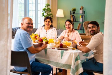 Gülümseyerek kameraya bakıyorum, neşeli Hintli aile, kıdemli ebeveynler ve genç yetişkin kız ve erkek çocuklar birlikte kahvaltı masasında oturuyorlar mutlu bir sabah anlarında.